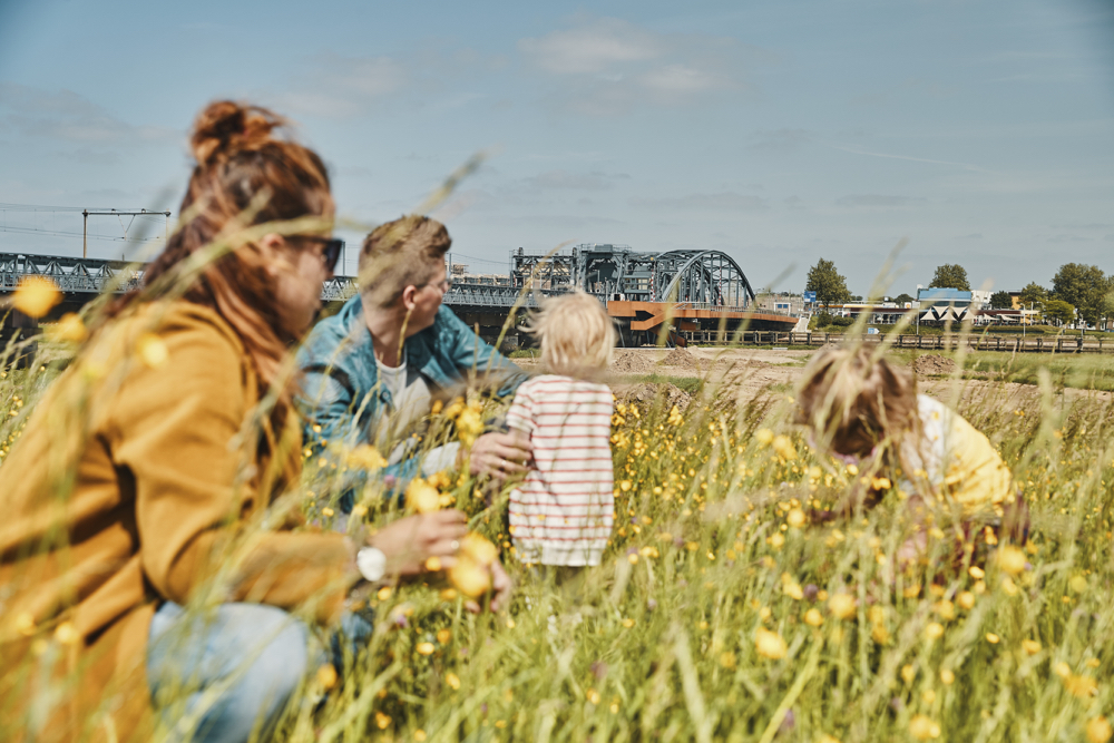 Een gezin met twee kindjes tussen de bloemen in het gras, met op de achtergrond de oude IJsselbrug