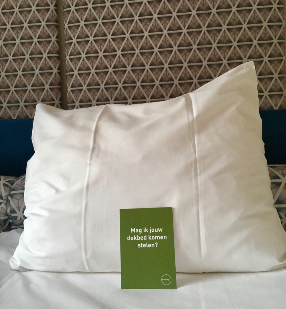 Een wit kussen op een wit dekbed met een groen kaartje met de tekst: mag ik je deken pakken