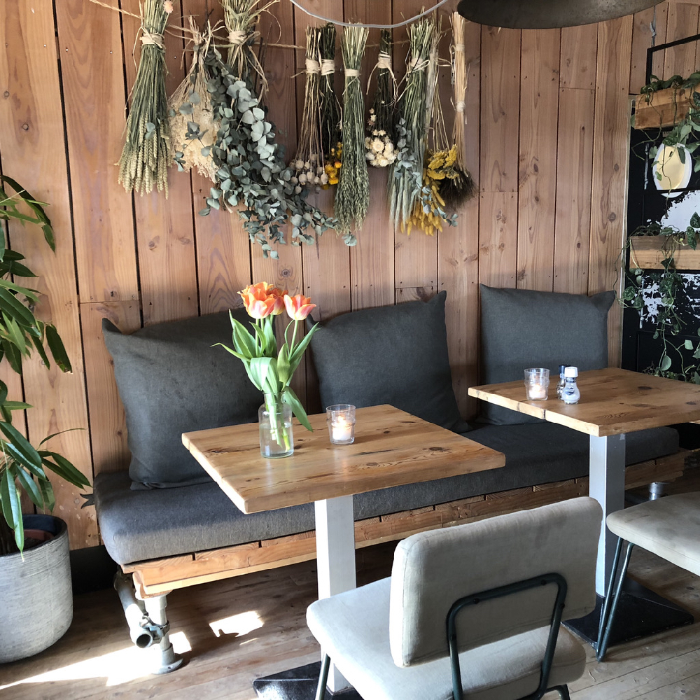 Bank met grijze kussens, gedroogde planten en bloemen aan de muur, twee tafeltjes met stoelen