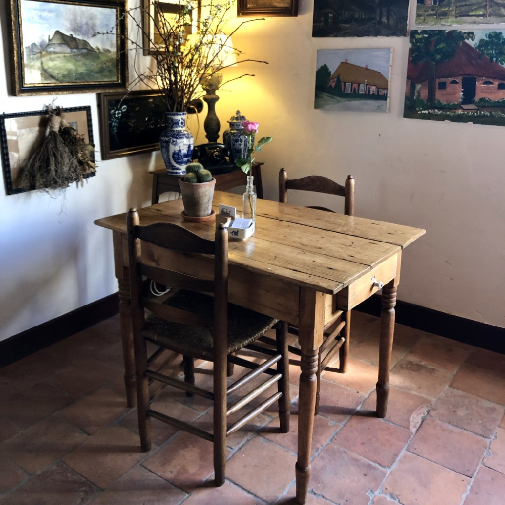 Twee houten stoeltjes aan een houten tafeltje met bloemen er op. Aan de muur hangen diverse schilderijen van boederijen