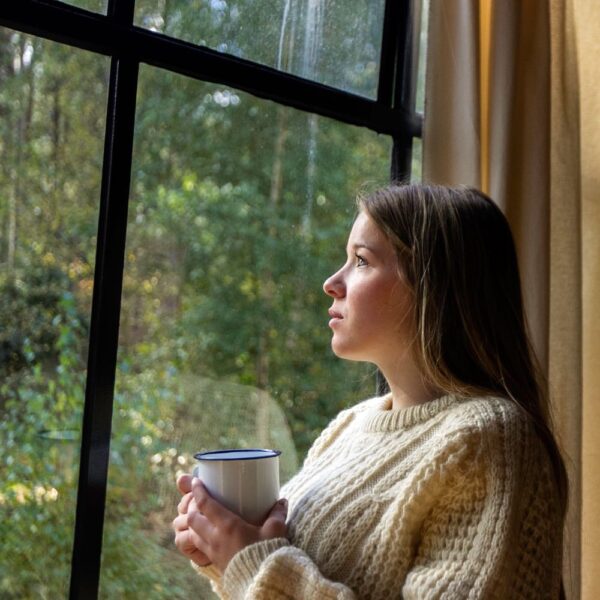 Jonge vrouw kijkt door het raam naar buiten met een kop koffie in haar handen