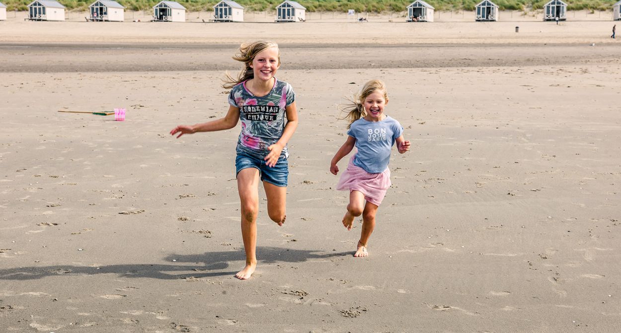 Zwei Mädchen, die am Strand laufen, mit Strandhäusern im Hintergrund. Der perfekte Ort für einen kinderfreundlichen Urlaub
