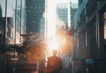 Oltre la sindrome da rientro — Quando a soffrire è il nostro legame con la natura Silhouette di un uomo in abito professionale che cammina da solo in una strada cittadina tra moderni grattacieli al tramonto, tenendo una valigetta.