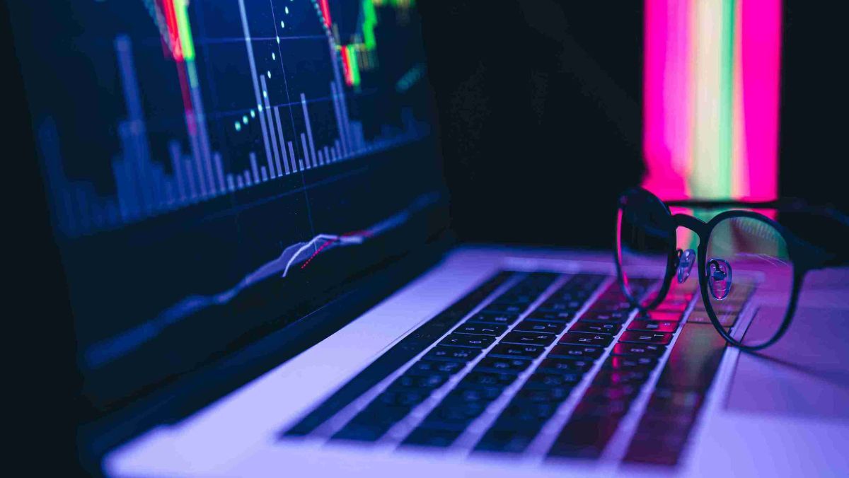 A laptop with stock charts on the screen and eyeglasses positioned on the keyboard