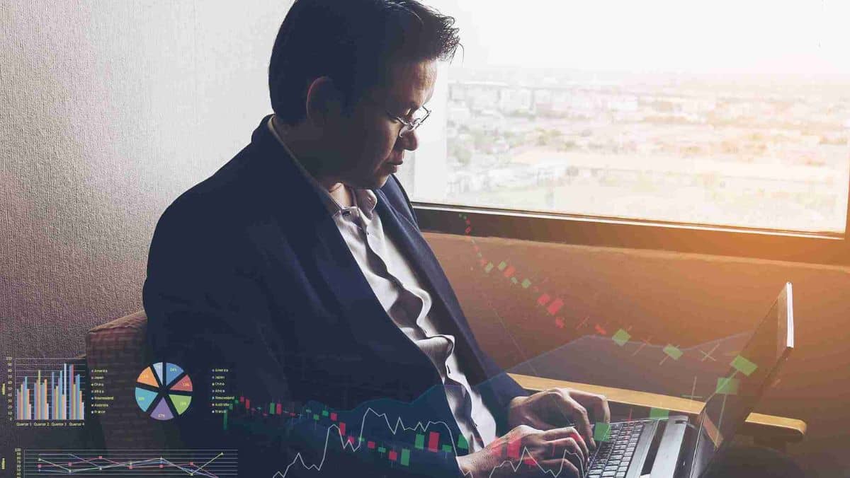 A man in formal attire carefully analyzing information on his laptop, with trading patterns visible in the background