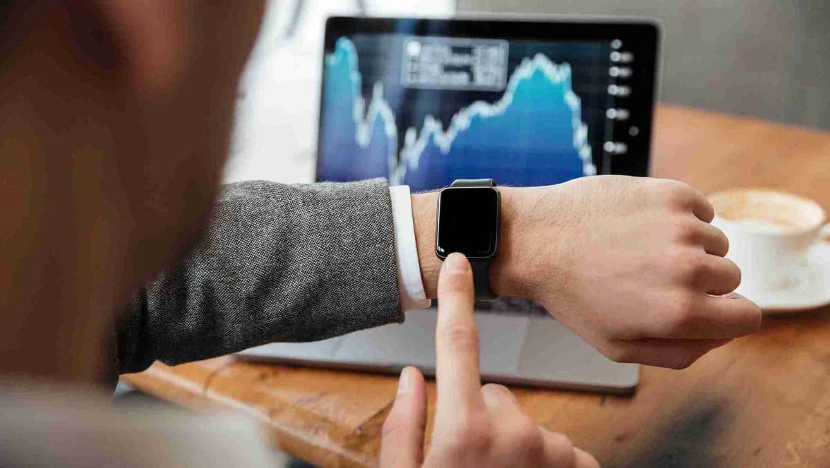 A man is looking and touching his wristwatch while an open laptop on the table shows a trading chart
