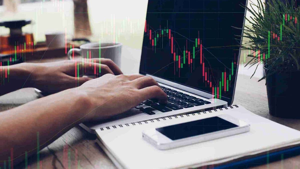 A man typing on a laptop next to a mobile phone, with floating candlestick patterns in the background
