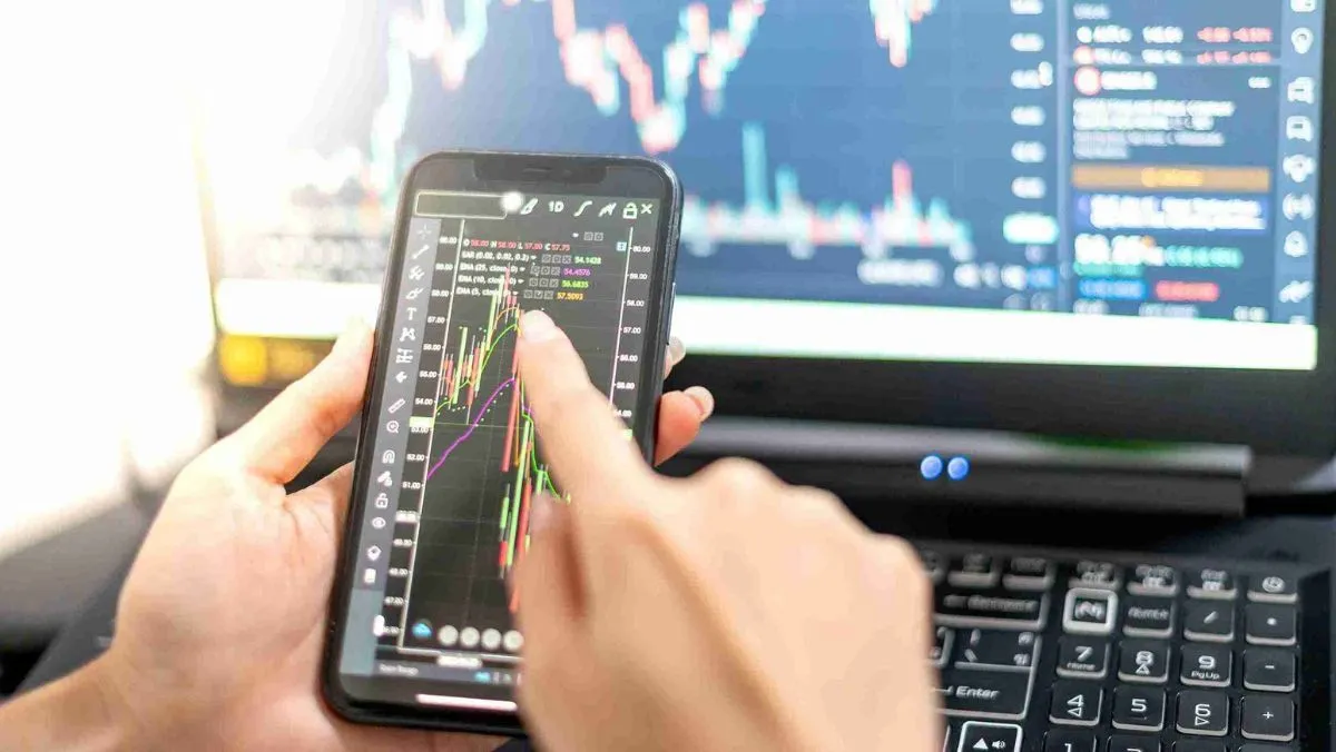 A person directs attention to a live trade on a mobile device while a computer on the table presents financial chart