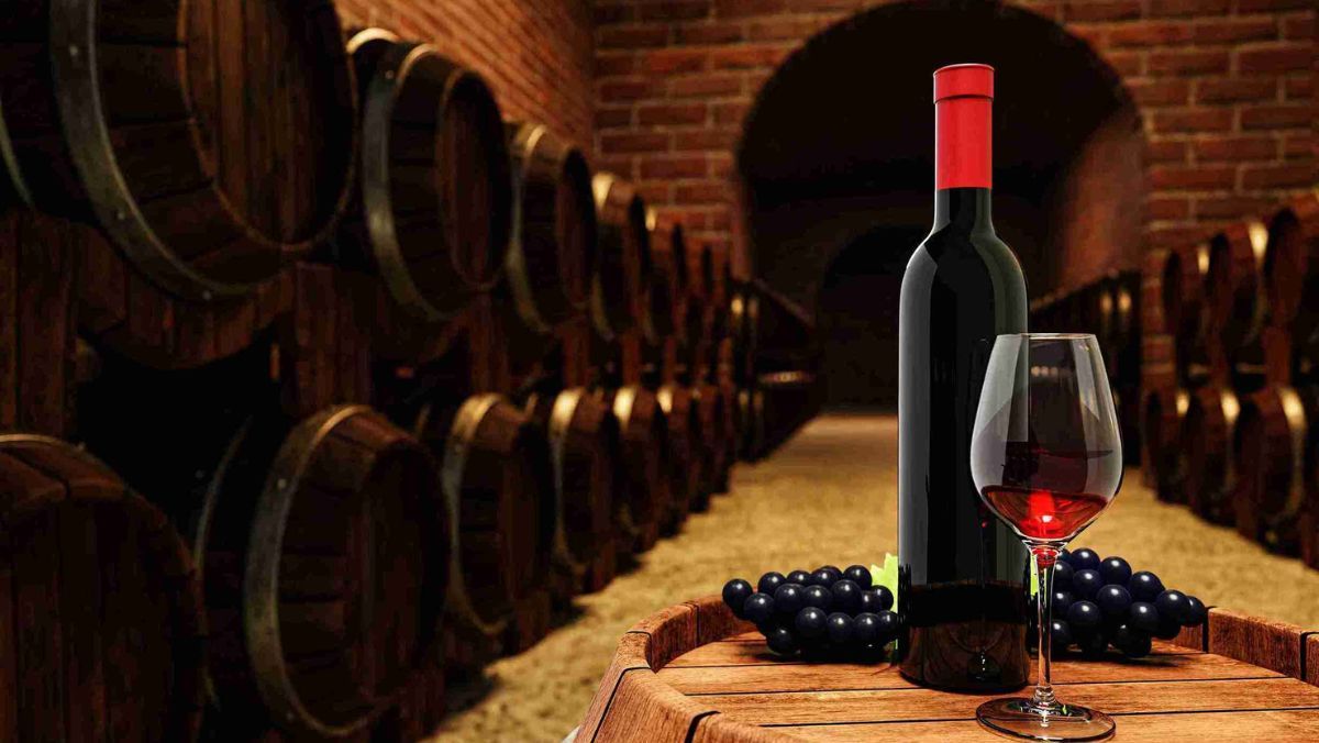 A red wine bottle and a clear glass containing red wine atop a wine fermentation tank surrounded by other fermentation tanks