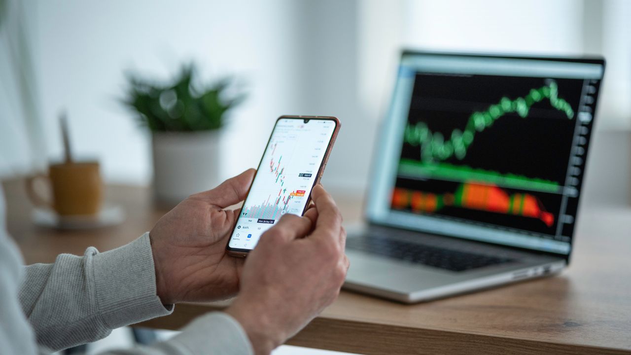 A person using a smartphone with stock charts on the screen and a laptop with trading graphs in the background
