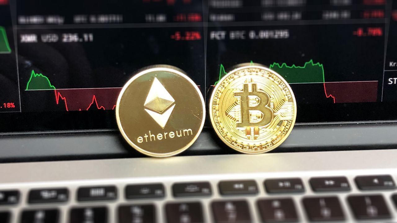 Physical coins representing Ethereum and Bitcoin placed on a laptop keyboard with stock market graphs in the background