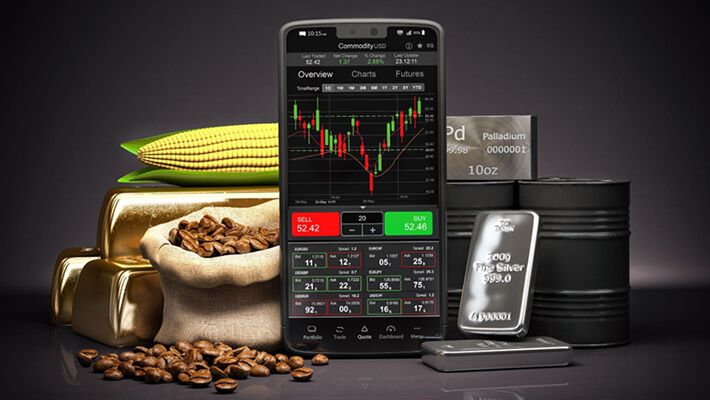 A smartphone displays commodity prices, surrounded by gold bars, coffee beans, corn, silver, and oil containers