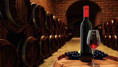 A red wine bottle and a clear glass containing red wine atop a wine fermentation tank surrounded by other fermentation tanks