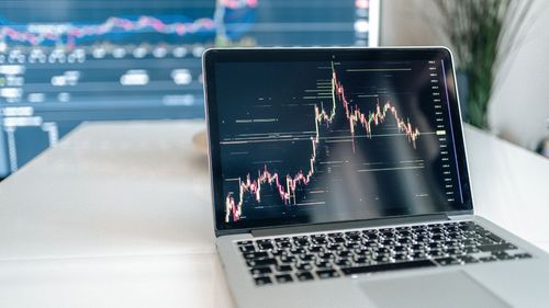 Open a laptop on a desk displaying stock market charts with a blurred background of trading monitors