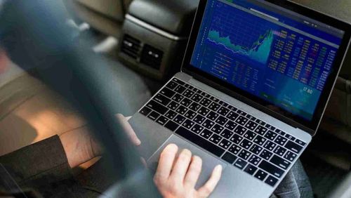 Positioned on a person's lap, a laptop is open, revealing a trading screen